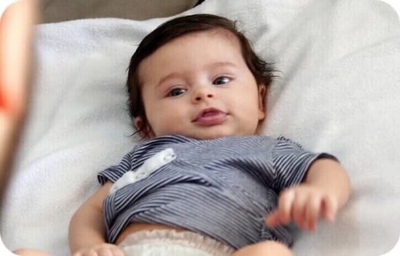 Photo of a baby with dark hair lying on a white pillow, gray striped onesie, playful expression