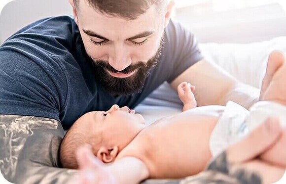 Photograph of smiling bearded man tenderly holding newborn on bed, soft light